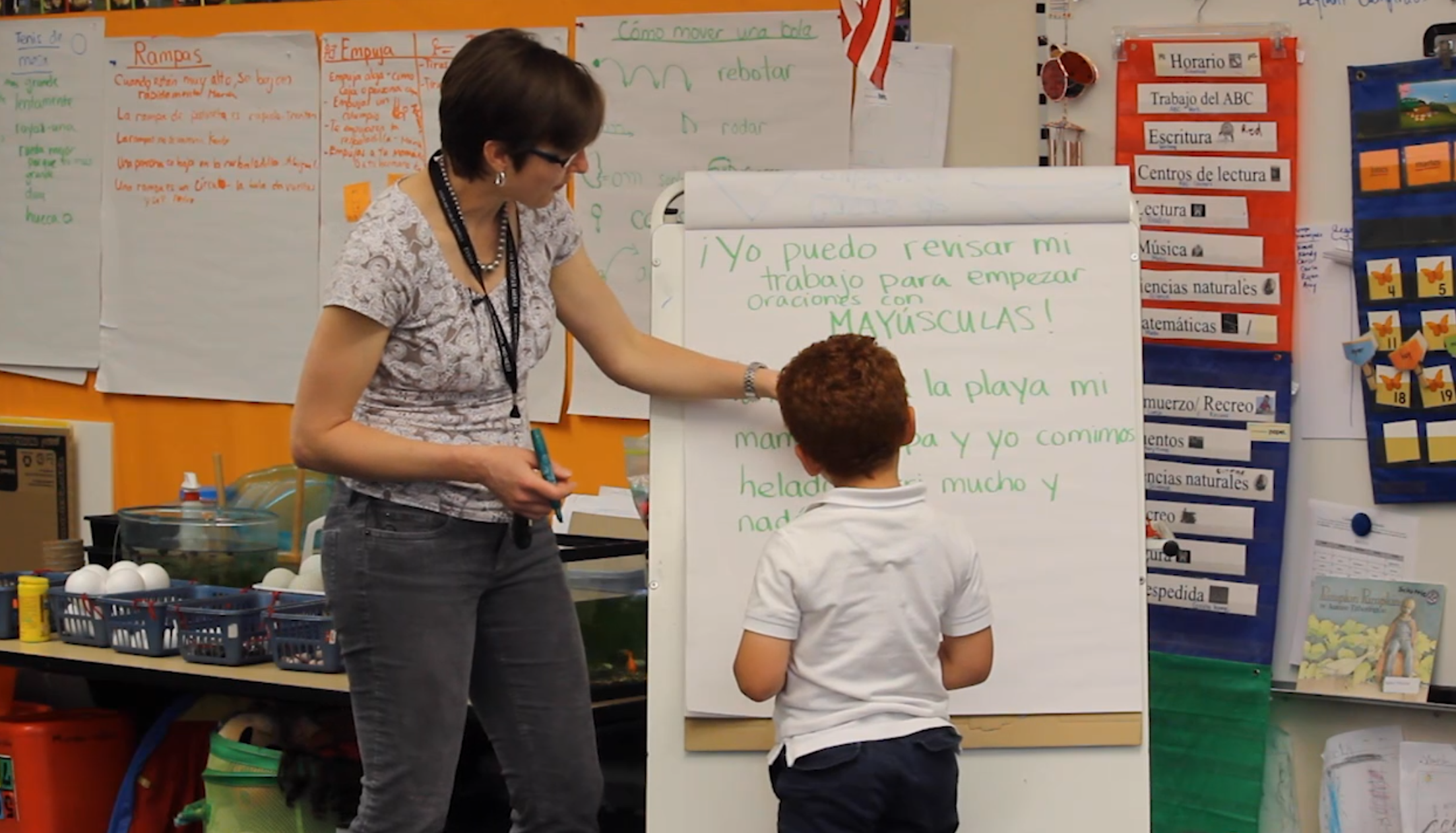 The image shows a teacher pointing to spanish words on a classroom display board while a child watches closely.